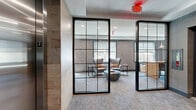 Located on the second floor of 975 Bonnel in Union, NJ, this thoughtfully designed common area serves as an elegant shared lounge space. The layout encompasses a welcoming foyer adjacent to elevator access, transitioning seamlessly into an open living area enclosed by striking floor-to-ceiling black-framed glass partition walls — a defining architectural feature that adds transparency and visual depth throughout.

The lounge area is furnished with plush wingback chairs accented by warm throw pillows, arranged around a sculptural stone coffee table, creating an inviting seating arrangement bathed in natural light from large exterior-facing windows. The windows offer views of the surrounding Union, NJ neighborhood, and are fitted with roller shades for adjustable light control.

The space also incorporates a dedicated work or dining area outfitted with wooden tables and chairs, pendant lighting overhead, and a wall-mounted television — making it functional for both relaxation and productivity. Textured geometric wallcoverings in muted grey tones add visual interest alongside warm wood accent panels, while plush patterned carpeting runs throughout, softening the overall aesthetic.

The foyer area features a wood credenza topped with decorative ceramics beneath a large round mirror, contributing to the polished, contemporary character of the space. Recessed lighting and clean white ceilings complete the refined, professional ambiance.