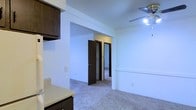 This single-floor residential space offers a straightforward and functional layout designed for comfortable everyday living. The interior is characterized by clean white walls and smooth ceilings that help reflect light throughout the space, contributing to a bright and open ambiance. A ceiling fan is installed in the main living area, providing both lighting and air circulation.

The living area features wall-to-wall carpeting in a neutral tone, creating a warm and inviting atmosphere. A partial wall or breakfast bar divides the kitchen area from the main living space, offering a sense of separation while maintaining an open feel. The kitchen is equipped with dark espresso-toned cabinetry and light-colored countertops, providing a classic contrast that anchors the space.

A central hallway connects the living area to the additional rooms, lined with dark wood interior doors that complement the kitchen cabinetry. Built-in shelving is visible within one of the doorways, suggesting dedicated storage or closet space. The consistent use of dark wood tones throughout the doors and cabinetry lends a cohesive and grounded aesthetic to the overall interior. The space presents a practical and well-organized floor plan suited for a variety of living arrangements.
