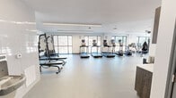 Located at 5102 Station Dr in Birmingham, AL, this well-appointed fitness center offers a spacious, single-floor layout designed to support a comprehensive workout experience. The space features a clean, modern aesthetic characterized by crisp white walls, smooth light-toned flooring, and a bright, open atmosphere enhanced by an abundance of natural light streaming through large floor-to-ceiling windows that frame outdoor views of the surrounding landscape.

The cardio zone is prominently featured along the window wall, housing a row of treadmills, elliptical trainers, stationary bikes, and a rowing machine — all equipped with individual digital displays. A dedicated strength and functional training area includes a multi-station cable and weight machine, a dumbbell rack with flat benches, and functional training accessories such as medicine balls, resistance equipment, and plyometric boxes. Linear LED lighting runs across the ceiling, providing consistent, even illumination throughout the space.

A built-in storage and amenity station along one wall includes open shelving and cabinetry with a countertop, a wall-mounted coat/towel hook rail, and a water fountain, adding practical convenience for users. The overall design balances utility and aesthetics, creating an inviting and fully equipped environment for fitness at any level.