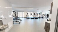 Located at 5102 Station Dr in Birmingham, AL, this well-appointed fitness center offers a spacious, single-floor layout designed to support a comprehensive workout experience. The space features a clean, modern aesthetic characterized by crisp white walls, smooth light-toned flooring, and a bright, open atmosphere enhanced by an abundance of natural light streaming through large floor-to-ceiling windows that frame outdoor views of the surrounding landscape.

The cardio zone is prominently featured along the window wall, housing a row of treadmills, elliptical trainers, stationary bikes, and a rowing machine — all equipped with individual digital displays. A dedicated strength and functional training area includes a multi-station cable and weight machine, a dumbbell rack with flat benches, and functional training accessories such as medicine balls, resistance equipment, and plyometric boxes. Linear LED lighting runs across the ceiling, providing consistent, even illumination throughout the space.

A built-in storage and amenity station along one wall includes open shelving and cabinetry with a countertop, a wall-mounted coat/towel hook rail, and a water fountain, adding practical convenience for users. The overall design balances utility and aesthetics, creating an inviting and fully equipped environment for fitness at any level.