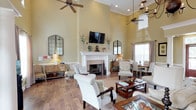 This single-story clubhouse offers an inviting and well-appointed community gathering space defined by dramatic soaring ceilings, warm hardwood flooring, and a cohesive neutral color palette throughout. The grand entrance greets visitors with rich wood French doors framed by classical columns, transom windows that draw in abundant natural light, and an elegant iron chandelier overhead. A welcoming reception area with a dark wood desk and comfortable seating provides a professional point of contact upon entry.

The expansive main lounge is a standout feature, anchored by a classic white-surround wood-burning fireplace topped with a mounted flat-screen television and decorative mantel accents. Multiple seating arrangements — including upholstered armchairs and sofas — create intimate conversation areas suited for relaxation or social gatherings. Ceiling fans are positioned throughout to ensure comfort, while arched and transom windows maintain a bright, airy atmosphere.

A dedicated dining area with a wooden table and upholstered chairs offers a more structured setting for meetings or casual meals. Adjacent to the main lounge, a private office space with a full-sized desk and filing storage provides a functional workspace. A Starbucks coffee station is also available, adding a convenient amenity for residents. Crown molding, decorative mirrors, and thoughtful furnishings complete the polished, community-focused ambiance of this clubhouse.