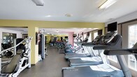This fitness center is a thoroughly equipped facility designed to support a wide variety of workout routines. The space is organized into distinct zones, each dedicated to a specific type of exercise, allowing for efficient use and easy navigation throughout.

The cardio zone features a row of treadmills and stationary bikes, positioned to take advantage of natural light streaming through large windows. Mounted flat-screen televisions provide entertainment during cardio sessions. Adjacent to this area, a selection of cable machines and multi-function strength stations offer users a comprehensive range of resistance training options.

The free weights section is well-stocked with a full rack of dumbbells spanning a broad range of weights, accompanied by adjustable benches and barbells for targeted strength work. Full-length mirrors line the walls of this area, enhancing the sense of space and allowing users to monitor their form during exercises.

Throughout the facility, durable rubber flooring provides safe, cushioned footing suitable for all types of training. The walls are painted in warm yellow and neutral tones, contributing to an energizing yet comfortable atmosphere. Overhead fluorescent lighting ensures the entire space is consistently and clearly illuminated. The layout flows openly between zones, making this fitness center a functional and welcoming environment for users of all fitness levels.