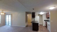 This single-floor residence offers a practical and comfortable layout designed for everyday living. The open-concept main area combines the living room and kitchen in one cohesive space, creating a sense of openness and ease of movement. The living area features wall-to-wall carpet in a warm, neutral tone, while the kitchen transitions to durable hardwood-style flooring, clearly delineating the two functional zones. The kitchen is equipped with white appliances, including a full-size refrigerator, and features dark-finished cabinetry that provides a striking contrast against the light walls. A freestanding dark-toned kitchen island anchors the space, offering additional counter and storage space. The textured ceilings add a classic residential character throughout. A doorway from the main living area leads into an adjacent room, visible in the background, which features carpeting and a ceiling fan, suggesting a bedroom or secondary living space. Neutral-toned walls, white baseboards, and recessed ventilation are consistent throughout, lending a clean and well-maintained aesthetic to the overall interior. The layout is straightforward and functional, making efficient use of the available space.