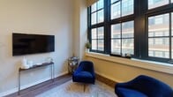 Located at 1 Market St in Camden, NJ, this single-floor unit showcases an impressive blend of industrial character and contemporary design. The living area is a standout, boasting soaring ceilings with exposed ductwork and oversized black-framed windows that flood the space with natural light and offer views of the surrounding urban streetscape. Rich hardwood-style flooring runs throughout the main living zone, grounding the open layout with warmth and continuity.

The living room is generously proportioned, accommodating a full seating arrangement with a sofa, accent chairs, and a glass coffee table, complemented by bold abstract artwork and pendant lighting that reinforce the loft-inspired aesthetic. A dedicated seating nook near the windows provides an ideal spot for relaxation or reading, bathed in daylight.

The kitchen is efficiently designed with a galley-style layout, featuring white cabinetry, light countertops, a full suite of white appliances including a refrigerator, electric range, and built-in microwave, along with a subway tile backsplash. Storage is well-appointed throughout.

The bathroom presents a clean, classic finish with white tile flooring, a pedestal sink, a mirrored medicine cabinet, and a shower-tub combination enclosed by a curtain. The neutral palette throughout the space creates a cohesive, move-in-ready environment suited for a variety of lifestyles.