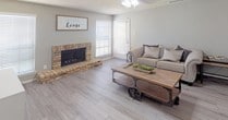 Located at 4450 Ridgemont Dr in Abilene, Texas, this single-floor home blends farmhouse charm with modern updates throughout. The living room serves as the heart of the space, showcasing a classic brick fireplace with a raised hearth, light wood-tone flooring, and neutral gray walls that create a warm, inviting atmosphere. A ceiling fan overhead and white plantation shutters on multiple windows allow for excellent light control and natural brightness. The kitchen features gray shaker-style cabinetry, a herringbone tile backsplash, stainless steel appliances, and a quartz countertop breakfast bar with rustic bar stool seating — ideal for casual dining or entertaining. The primary bedroom is generously proportioned, offering direct outdoor access through a set of French doors, a ceiling fan, and continuity of the wood-tone flooring. An ensuite bathroom provides a dual-sink vanity with quartz countertops, navy blue cabinetry, a large framed mirror, and well-appointed storage. A dedicated workspace area is also visible within the layout, adding functional versatility to the home. Throughout, the consistent use of updated flooring, neutral tones, and modern fixtures contributes to a cohesive, move-in-ready aesthetic.