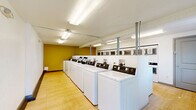 Located at 645 Wolff St in Denver, Colorado, this dedicated laundry facility offers a well-organized, single-floor space designed for efficient, shared use. The room accommodates a substantial row of top-loading washing machines lined along one wall, complemented by a separate bank of dryers, providing ample capacity for a multi-unit residential building. The machines feature digital control panels, indicating modern, user-friendly coin-operated or card-based operation. The floor is finished with light wood-toned vinyl tile, contributing a clean and practical aesthetic throughout the space. Walls are painted in a combination of crisp white and a warm mustard-yellow accent, adding a touch of color to the utilitarian environment. Overhead, fluorescent lighting fixtures provide bright, even illumination across the entire room. Structural support columns are positioned within the space, and exposed utility conduits and venting run along the ceiling. A row of windowed storage compartments or lockers is mounted along one wall, offering convenient space for residents. Natural light filters in through a window on one end, and a keyed exterior door provides secure, direct access. The layout ensures a comfortable, open walkway alongside the machines, making the facility easy to navigate during use.