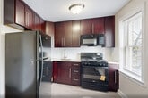 Located at 1127 John F. Kennedy Blvd in Bayonne, NJ, this single-floor, 2-bedroom, 1-bathroom residence offers a practical and comfortable layout with a clean, neutral aesthetic throughout. Rich, dark hardwood floors extend across the living areas and bedrooms, lending warmth and character to the space. The living room is well-proportioned and bright, featuring a ceiling fan and multiple windows that invite ample natural light, complemented by baseboard heating units and a wall-mounted AC unit for year-round comfort. The kitchen stands out with deep mahogany-toned cabinetry extending to the ceiling, light-toned tile flooring, a window for natural light, and a full suite of black appliances including a refrigerator, range, and over-the-range microwave. Adjacent pantry storage adds functional convenience. The two bedrooms are calm and neutral, with hardwood floors, white trim, and windows fitted with blinds offering neighborhood views. The primary bedroom is a comfortable size and features access to multiple closets — a notable storage advantage, with four closets distributed throughout the unit. The bathroom is clean and modern, showcasing white subway tile in the tub/shower surround, a glass shower door, a dark wood vanity with a white countertop, and a window for ventilation. A dedicated entrance and hallway complete the thoughtfully organized floor plan.
