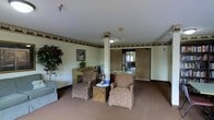 This single-floor space presents a traditionally styled common lounge area designed to provide a comfortable and welcoming environment. The room features neutral-toned carpeting throughout, which adds warmth underfoot and contributes to the overall cozy atmosphere. White ceilings are accented by a decorative wallpaper border that runs along the upper perimeter of the walls, adding a classic, ornamental touch to the interior.

A central support column serves as a defining architectural element, visually dividing the space into distinct seating zones. Floral-patterned armchairs are arranged around a small ornate side table, creating an intimate conversation area. A decorative arrangement of fresh flowers sits atop the table, adding a lively focal point. Additional seating, including a light blue chair, is positioned near a large indoor plant that introduces a natural element to the room.

Along one wall, a wooden upright piano is visible, suggesting the space may serve a social or recreational function. Framed artwork adorns the walls. Toward the rear of the space, wooden doors with sidelights provide access to adjacent areas, while overhead lighting fixtures illuminate the room evenly, reinforcing the space's practical and inviting character.