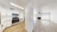 This single-floor residence offers a clean, modern aesthetic defined by a consistent palette of white walls, crisp trim, and wood-look tile flooring that runs throughout the entire space. The open-concept living area is generously proportioned, anchored by a fireplace set against a painted white brick accent wall, and fitted with a ceiling fan overhead. Natural light filters in through blinded windows, enhancing the bright and airy atmosphere. A pass-through counter connects the living area to the kitchen, which features white shaker-style cabinetry with matte black hardware, white quartz countertops, and a full suite of stainless steel appliances including a range, over-the-range microwave, and built-in lighting. The bedrooms continue the home's cohesive design, each showcasing the white brick accent walls, ceiling fans, and windows that invite natural light. At least one bedroom includes a walk-in closet, providing practical storage. The bathroom is well-appointed with a white vanity topped with a solid surface countertop, a large mirror, brushed gold hardware accents, and a painted brick wall that adds texture and character. The shower area features clean, bright surrounds and wood-look tile flooring, maintaining a unified look across the home.
