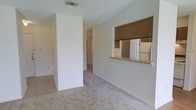 This single-story space showcases a bright, open layout designed for functional living. The main living area is finished with neutral-toned carpet and features vaulted ceilings that lend an airy sense of height and spaciousness. White walls throughout create a clean, minimalist backdrop that maximizes the natural light filtering into the space.

A defining architectural feature is the pass-through window connecting the living area to the kitchen, topped with warm-toned wood cabinetry above. This design element maintains an open, connected feel between the two areas while providing a convenient serving or interaction point. The kitchen is visible through the pass-through and includes white cabinetry and a refrigerator.

A structural half-wall or column adds subtle definition between zones without fully enclosing the space, contributing to the open-plan feel. Tile flooring is visible near the transition areas, complementing the carpeted living space. A hallway leads further into the unit toward additional rooms, suggesting a practical, well-organized floor plan. The HVAC vent integrated into the ceiling indicates a centralized climate control system throughout the space.