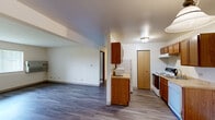 This single-floor unit offers a practical and efficiently designed layout across two bedrooms, one bathroom, and a combined living, dining, and kitchen area. The open-concept main living space features gray hardwood-style flooring that runs throughout, white walls, and a wall-mounted air conditioning unit. A large mirror panel in the dining area visually expands the space, while a pendant light fixture adds a decorative touch overhead.

The kitchen is outfitted with warm-toned wood cabinetry on both upper and lower sections, a white refrigerator, a white electric range with dishwasher, and a single-basin sink. A separate pantry closet provides additional storage.

The primary bedroom is generously sized and features hardwood-style flooring, a window with vertical blinds offering outdoor views of greenery, and a wall-mounted A/C unit. The second bedroom is carpeted with a window that also overlooks outdoor greenery, lending a bright, airy feel.

The bathroom includes a full tub-shower combination with a curtain rod, a wood-toned vanity with a single sink, and a mirror above. The overall design is clean and functional throughout.