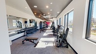 Located at 1300 5th Avenue South in Birmingham, Alabama, this fitness center offers a thoughtfully designed exercise space that balances functionality with modern aesthetics. The single-floor layout provides an open, elongated floor plan that accommodates a wide variety of workout zones without feeling crowded.

The space is finished with neutral gray carpet flooring and crisp white walls, accented by a bold pink feature wall that adds a vibrant pop of color. Large black-framed windows line the perimeter, flooding the interior with abundant natural light and offering views of the surrounding Birmingham cityscape. Oversized ceiling fans with a striking blade design are mounted overhead, complementing the recessed lighting to ensure consistent air circulation and illumination throughout.

The strength training area is prominently featured, with an extensive dumbbell rack spanning a full wall, supported by adjustable benches and weight machines suited for a variety of exercises. A dedicated power rack with plate storage anchors the free weights section, while cable machines and specialty strength equipment round out the resistance training offerings. Mirrors along one wall enhance the sense of space and provide visual feedback during workouts.

The cardio and functional training zones include rowing-style machines, stationary bikes, and open floor space for stretching or dynamic movement. Vibrant wall art adds personality to the environment, creating an energizing atmosphere. A wall-mounted television adds convenience for entertainment during training sessions.