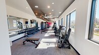Located at 1300 5th Avenue South in Birmingham, Alabama, this fitness center offers a thoughtfully designed exercise space that balances functionality with modern aesthetics. The single-floor layout provides an open, elongated floor plan that accommodates a wide variety of workout zones without feeling crowded.

The space is finished with neutral gray carpet flooring and crisp white walls, accented by a bold pink feature wall that adds a vibrant pop of color. Large black-framed windows line the perimeter, flooding the interior with abundant natural light and offering views of the surrounding Birmingham cityscape. Oversized ceiling fans with a striking blade design are mounted overhead, complementing the recessed lighting to ensure consistent air circulation and illumination throughout.

The strength training area is prominently featured, with an extensive dumbbell rack spanning a full wall, supported by adjustable benches and weight machines suited for a variety of exercises. A dedicated power rack with plate storage anchors the free weights section, while cable machines and specialty strength equipment round out the resistance training offerings. Mirrors along one wall enhance the sense of space and provide visual feedback during workouts.

The cardio and functional training zones include rowing-style machines, stationary bikes, and open floor space for stretching or dynamic movement. Vibrant wall art adds personality to the environment, creating an energizing atmosphere. A wall-mounted television adds convenience for entertainment during training sessions.