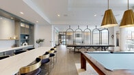 Located on 12th Ave S in Minneapolis, MN, this single-floor space combines a fully equipped kitchen and an open living room into one cohesive, thoughtfully designed layout. The kitchen area is anchored by a striking oversized island with a white marble-style countertop and a brushed gold base, surrounded by sleek black-and-gold bar seating. Custom sage-blue cabinetry lines the walls, complemented by white subway tile, white quartz countertops, open display shelving, a stainless steel sink, a beverage refrigerator, and a wall-mounted television — creating a functional yet refined culinary space. A cozy nook with upholstered banquette seating is tucked nearby, adding an intimate gathering spot. The living area transitions seamlessly from the kitchen, featuring wide-plank hardwood floors throughout, a built-in linear fireplace framed by warm wood shelving and display niches, and a wall-mounted screen. Floor-to-ceiling sliding glass doors flood the room with natural light and open directly to an outdoor terrace with city views, enhancing the sense of openness. Arched architectural glass partitions and bold black-framed doors add refined contrast. The overall ambiance is modern and upscale, balancing clean lines with warm, inviting textures throughout.
