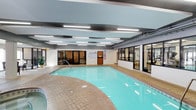 This single-floor indoor recreation facility is designed to provide a comprehensive wellness experience. At its heart is a generously sized indoor swimming pool with clear, turquoise water, surrounded by a wide brick-patterned tile deck that offers ample space for entry and movement around the pool. A hot tub is conveniently positioned adjacent to the pool, providing a relaxation option alongside aquatic activities. The ceiling features a modern suspended panel system with integrated fluorescent lighting, ensuring consistent, bright illumination throughout the space regardless of the time of day. The structural beams and ductwork are incorporated into the ceiling design, giving the space an open, functional aesthetic. Large glass panel windows and doors divide the pool area from an adjoining fitness center, which is visible and accessible directly from the pool deck. The fitness center appears equipped with cardio and strength training equipment. Safety and informational signage is posted near the pool entrance, reflecting a well-managed and regulated environment. The overall ambiance is clean, practical, and purposefully designed for active recreation and relaxation.