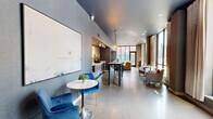 Located at 18 W 15th St in Minneapolis, MN, this contemporary single-floor space combines a fully equipped kitchen and an open living area into one cohesive, modern environment. The interior design is defined by polished concrete floors, deep blue-grey ceilings, and textured wall treatments that give the space an urban, sophisticated character throughout.

The kitchen runs along one wall and is outfitted with sleek grey cabinetry, bright white countertops, a stainless steel sink, a wine cooler, a beverage station, and under-counter appliances. A large piece of abstract wall art adds a creative focal point above the counters, while a wood-paneled accent wall lends warmth. A high-top island with bar seating sits at the center of the space, illuminated by stylish pendant lights, and serves as both a dining and gathering area.

The living area is anchored by a generous sectional sofa and complemented by a sleek round coffee table and accent chairs. Floor-to-ceiling black-framed windows wrap the lounge corner, flooding the space with natural light and offering open views of the surrounding Minneapolis streetscape. Sheer curtains frame the windows elegantly. A set of glass-panel doors with black steel frames provides a bold, transparent entry point, maintaining visual connectivity and allowing light to flow freely throughout the entire space.