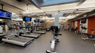 This single-floor fitness center offers a comprehensive workout environment thoughtfully organized across several distinct zones. The main cardio area is generously proportioned, accommodating a long row of treadmills, elliptical machines, and stationary bikes — including both upright and recumbent models — all positioned for easy use and circulation. Multiple wall-mounted TVs throughout keep members entertained during their sessions. A dedicated strength training section houses a variety of cable machines and resistance equipment, providing ample options for full-body conditioning. A separate, more intimate free weights room features a cable crossover station, adjustable benches, a stability ball, and a wall-mounted TV, making it well-suited for focused or personal training sessions. A small lounge area with a red sofa, patterned accent chairs, and a round marble-top coffee table offers a comfortable space to rest or wait. Throughout the facility, a striking reclaimed wood feature wall with a chevron pattern adds warmth and visual character, contrasting with the industrial-style exposed ductwork and concrete ceilings. Durable rubber flooring, pendant lighting, and full-length mirrors complete the practical yet stylish design of this well-rounded fitness space.