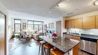 This contemporary single-floor residence is thoughtfully designed with an open-concept layout that seamlessly connects the kitchen, dining, and living areas. The kitchen is a standout, featuring warm medium-tone wood cabinetry, dark granite countertops, a classic subway tile backsplash, and a full suite of stainless steel appliances including a double oven, dishwasher, microwave, and refrigerator. A central island with a matching granite surface and bar seating serves as both a functional workspace and a casual dining hub. Track lighting overhead provides ample illumination throughout the cooking and gathering space. Hardwood flooring flows through the open living area, which is bathed in natural light from a wall of floor-to-ceiling windows with views of the surrounding urban landscape. The living room offers generous space for comfortable seating and entertainment. The bedroom is carpeted and serene, featuring a large window with outdoor views, a walk-in closet with open shelving and hanging storage, and a warm, understated ambiance. The full bathroom is finished with large dark-format floor tiles, a single-sink vanity with wood cabinetry, a wide mirror, and a spacious glass-enclosed walk-in shower with grey subway tile surround.