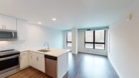 This single-floor residence showcases a clean, contemporary design with an open-concept layout that seamlessly connects the kitchen and living area. The kitchen is a standout feature, equipped with crisp white cabinetry extending to the ceiling, a classic white subway tile backsplash, and a suite of stainless steel appliances including a French door refrigerator, built-in microwave, and range. A generous quartz countertop island with an integrated sink serves as both a functional workspace and a natural gathering point. Rich, dark-toned hardwood flooring runs throughout the main living areas, providing a warm contrast to the bright white walls and recessed lighting overhead. Large, floor-to-ceiling windows in the living area flood the space with natural light and offer views of the surrounding streetscape. The bedroom is a well-proportioned, versatile space featuring the same hardwood flooring and exposed ductwork that adds an industrial accent. The bathroom is finished to a high standard, with large-format light tile flooring, a dark espresso vanity with a white countertop, and a backlit LED mirror that lends a polished, modern feel. Throughout the unit, the design balances functionality with a fresh, modern aesthetic.