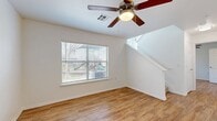 Located in Humble, Houston, TX, this two-story residence offers three bedrooms and two bathrooms across a well-utilized layout spread over two floors. The main level, which comprises the larger portion of the home, features an open-concept living area finished with warm wood-style laminate flooring throughout. A ceiling fan with integrated lighting anchors the living room, which flows seamlessly toward the kitchen via a peninsula counter with granite-style countertops. The kitchen is equipped with rich dark-wood cabinetry, black appliances including a refrigerator and microwave, and track lighting overhead. A dedicated laundry area on the main floor includes washer and dryer hookups and is outfitted with a utility shelf. The upper floor hosts the bedrooms, each featuring plush carpeting, ceiling fans, and windows with blinds that allow natural light. The spacious primary bedroom offers a comfortable retreat with access to the hallway and nearby closets. Both bathrooms are finished with dark wood vanities, granite-style countertops, large mirrors, and wood-style flooring, lending a consistent and polished aesthetic throughout. Clean white walls, practical storage, and a cohesive design make this a functional and well-appointed home.
