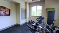 This single-floor fitness space is designed to support focused cardio training, centered around a lineup of stationary cycling bikes arranged in a row along one wall. The room features an arched window that frames views of lush outdoor greenery, including palm trees, allowing natural light to brighten the workout environment. The walls are finished in a neutral beige tone, complemented by dark patterned carpet flooring that provides cushioning underfoot. A ceiling fan overhead aids in air circulation during intense sessions. A louvered door with a matching side panel offers access to an adjacent area, adding functional flow to the layout. The overall ambiance is clean and utilitarian, with an emphasis on practicality and comfort for cyclists of all levels.