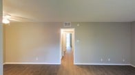 This single-floor space presents a clean, minimalist aesthetic defined by neutral gray walls and warm hardwood-style flooring that runs consistently throughout. The main room is open and unfurnished, offering a blank canvas with a sense of generous width and depth. White baseboards provide crisp contrast against the gray walls, contributing to a tidy, well-maintained appearance. Ceiling-mounted HVAC vents are visible, indicating a central climate control system throughout the unit. A centrally positioned doorway leads into a connecting hallway, which opens to additional rooms beyond. Natural light filters through what appears to be a large window or sliding glass door at the far end of the hallway, brightening the interior and adding depth to the layout. A smoke or CO detector is mounted on the wall near the hallway entrance. The overall ambiance is neutral and move-in ready, with a simple, functional design that lends itself well to a variety of uses. The consistent flooring and color palette across visible spaces create a cohesive, unified feel throughout the interior.