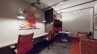 This thoughtfully designed single-floor workspace blends productivity and comfort through a cohesive palette of bold red, deep walnut wood tones, and neutral grays. The space is anchored by a dramatic built-in lounge nook, clad in rich dark wood paneling with vibrant red upholstered bench seating and decorative throw pillows, creating an intimate setting ideal for relaxed collaboration or quiet focus. Overhead, a cluster of pendant lights illuminates the nook with a warm glow.

Adjacent to the lounge area, a long wall-mounted workstation desk runs the length of one side, accompanied by ergonomic red mesh chairs, making it well-suited for individual work sessions. A bold abstract artwork in red, orange, and white tones dominates the wall above the desk, adding a striking visual focal point. A second inspirational text-based artwork further enhances the creative atmosphere.

A small round café-style table with contemporary chairs provides an additional collaborative seating option. Patterned area rugs layer over solid red carpet flooring, defining zones within the open layout. A service counter with dark cabinetry and a multifunction printer supports day-to-day office needs. The ceiling features integrated lighting tracks and recessed fixtures, ensuring the space remains well-lit throughout. Overall, the space exudes a vibrant, creative, and professional ambiance.