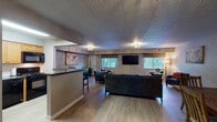 This single-floor space is thoughtfully laid out with an open-concept design that connects the main living area, dining space, and kitchen into a cohesive, functional environment. Throughout, light wood-tone flooring provides a warm and consistent foundation, complemented by neutral-toned walls and textured tile ceilings.

The spacious living area is furnished with multiple dark upholstered sofas and accent chairs, centered around a large wall-mounted flat-screen television flanked by two windows dressed with patterned roman shades and horizontal blinds. Natural light filters in generously, enhancing the welcoming atmosphere. A world map artwork serves as a prominent decorative focal point, and an ornate flush-mount chandelier adds a refined touch overhead.

A dedicated seating nook features two leather accent chairs arranged around a small bistro-style side table, creating an intimate conversation area. Adjacent to this is a compact dining space with a rustic wood dining table and matching slatted chairs, accented by a decorative wall clock.

The kitchen is equipped with light wood cabinetry, black appliances including a refrigerator, range, and microwave, a white subway tile backsplash, and dark granite countertops extending to a peninsula with bar seating, seamlessly connecting the kitchen to the living area.