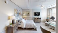 Located at 82 E Tennessee Ave in Oak Ridge, TN, this efficiently designed one-bedroom, one-bathroom unit makes excellent use of its compact footprint across a single floor. The living area is bathed in natural light streaming through large windows fitted with horizontal blinds, offering views of the surrounding cityscape. A built-in shelving unit along one wall provides both display and storage space, complementing the warm light wood flooring that runs throughout the unit.

The generously proportioned bedroom serves as the centerpiece of the layout, comfortably accommodating a full bed, dresser, and wall-mounted television. Soft, neutral wall tones and a ceiling fan contribute to a calm, restful atmosphere, while colorful artwork adds a personalized touch.

The kitchen is thoughtfully appointed with white upper cabinetry, blue lower cabinetry, granite-look countertops, a stainless steel refrigerator, and in-unit washer — maximizing functionality in a compact space. Louvered closet doors provide additional storage access nearby.

The bathroom features wood-look flooring, a white vanity with mirror, and a frosted glass window that draws in soft diffused light while maintaining privacy. Clean, neutral finishes throughout the unit create a cohesive and inviting aesthetic well-suited for comfortable everyday living.