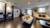 This contemporary single-floor amenity space is thoughtfully designed to blend comfort, style, and functionality in a cohesive modern aesthetic. Rich, dark hardwood-style flooring runs throughout, creating a striking contrast against the crisp white walls and recessed lighting overhead.

The lounge area serves as the social centerpiece, furnished with sleek white leather sofas arranged around a bold, dark built-in media wall featuring open shelving and a flat-screen television. Large glass entry doors flood the space with natural light and offer views of a lush outdoor area, enhancing the open, welcoming atmosphere.

Adjacent to the lounge, the business center provides individual workstations set within dark architectural column structures, each outfitted with ergonomic white chairs — ideal for focused, professional work. Glass-partitioned walls define the area while maintaining visual openness.

A dedicated conference room accommodates group meetings around a large oval conference table surrounded by modern task chairs. The room is equipped with a printer station, snack and beverage vending machines, and an ATM, offering a full suite of conveniences. Striped curtains frame a window, softening the space with natural light. Colorful abstract artwork adorns the walls throughout, adding creative energy to the professional environment.