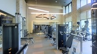 This contemporary fitness center is designed to deliver a premium workout experience within a thoughtfully appointed space. Soaring double-height ceilings create an open, airy atmosphere, while large floor-to-ceiling windows along one wall flood the interior with natural light and offer views of the surrounding greenery. Sleek linear pendant lighting fixtures hang overhead, complementing the neutral, modern color palette of warm whites and soft grays throughout.

The facility is comprehensively equipped with a wide array of strength-training machines, free weights, and benches arranged in an organized, accessible layout. A full dumbbell rack lines one wall, while multiple cable and resistance machines are positioned across the floor to accommodate a variety of workout routines. Cardio equipment is also present, ensuring a well-rounded fitness experience.

Large decorative wall art featuring bold, abstract brushstroke designs adds a sophisticated aesthetic touch, while floor-to-ceiling mirrors along the back wall visually expand the space and allow users to monitor their form. The flooring features a durable, neutral-toned surface suited for high-traffic use. Natural stone or brick accent walls contribute texture and warmth, grounding the modern design with an approachable, upscale character throughout.