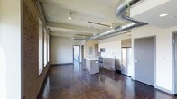 This single-floor space in Dallas showcases a compelling blend of industrial and contemporary design. The open-concept layout allows the living and kitchen areas to flow seamlessly together, creating a spacious and versatile environment. Exposed silver ductwork runs prominently across the ceiling, lending an industrial character that is complemented by recessed lighting and a concrete ceiling with a raw, unfinished aesthetic.

The kitchen is a standout feature, equipped with stainless steel appliances including a full-size refrigerator, built-in oven, and microwave. Cabinetry combines crisp white upper cabinets with warm wood-toned lower cabinets and a matching kitchen island, which provides additional prep space and casual seating. A sleek countertop and single-basin sink add to the clean, modern finish.

The flooring throughout is polished dark concrete, adding depth and a sophisticated industrial edge to the space. A row of large windows along one wall invites natural light, enhancing the open feel. A corridor visible in the background leads to additional areas of the unit, suggesting a thoughtful and functional floor plan overall.