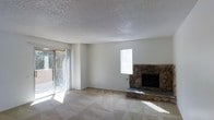 This single-story space centers around a spacious living area that combines functional design with inviting character. The room's most prominent feature is a floor-to-ceiling stone fireplace, constructed from natural stacked rock, which serves as a striking focal point along one wall. Its earthy, textured appearance adds a rustic warmth that contrasts with the otherwise neutral palette of the room.

Natural light is a defining characteristic of the living area. A large sliding glass door along one wall opens to an outdoor space visible through the glass, allowing ample sunlight to pour across the carpeted floor. An additional window on the adjacent wall further enhances the brightness and airiness of the room. The carpet features a subtle geometric pattern, adding a understated visual texture to the floor.

The walls are finished in a clean, neutral white tone, complementing the textured popcorn ceiling overhead. The overall design is open and uncluttered, providing a versatile canvas for various furnishing arrangements. The combination of the stone fireplace, abundant natural light, and simple finishes gives the space a comfortable, lived-in character well-suited for relaxed everyday living.