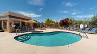 Located at 501 W Zia Rd in Santa Fe, New Mexico, this outdoor recreational area offers a well-appointed poolside retreat designed for relaxation and leisure. At the heart of the space is a freeform swimming pool with clear turquoise water, accessible via dual stainless steel entry rails and surrounded by a generous concrete deck that provides ample room for sunbathing and circulation.

The pool deck is furnished with multiple blue lounge chairs arranged to maximize sun exposure, making it an ideal spot for unwinding under New Mexico's abundant sunshine. A dedicated seating area features a shaded dining table with a tan market umbrella, surrounded by additional chairs — perfect for socializing or enjoying refreshments outdoors. A pergola structure nearby adds architectural interest and provides a shaded alternative to open-air seating.

The entire area is enclosed by a wrought iron fence, ensuring both safety and a sense of privacy. The perimeter is richly landscaped with mature evergreen trees, ornamental shrubs, and a striking red-leafed tree that adds vibrant seasonal color. The surrounding adobe-style buildings are visible in the background, grounding the space in the distinctive Southwestern architectural aesthetic of Santa Fe. Abundant natural light and open sky views complete this inviting outdoor amenity.