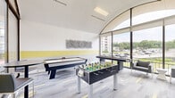 Located in College Park, Maryland, this dedicated game room is a well-appointed recreational space designed for social engagement and leisure. The room's most striking architectural feature is its soaring arched ceiling paired with expansive floor-to-ceiling windows that flood the interior with abundant natural light and offer open views of the surrounding greenery and streetscape.

The space is outfitted with a diverse selection of gaming equipment, including a foosball table, an air hockey table, a shuffleboard table, and a standalone arcade machine, providing entertainment options for a wide range of interests. Comfortable seating, including a pair of upholstered lounge chairs and tall bistro-style tables with bar stools, offers spaces to relax and socialize between games.

The interior design reflects a clean, modern aesthetic. Walls are finished in crisp white with a bold yellow horizontal accent band, echoing the yellow highlights woven into the multi-toned carpet tiles below. Abstract wall art adds a creative touch to the décor. The combination of thoughtful design, ample natural light, and a well-rounded selection of games makes this room a dynamic and inviting communal amenity.