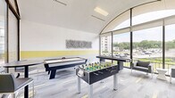 Located in College Park, Maryland, this dedicated game room is a well-appointed recreational space designed for social engagement and leisure. The room's most striking architectural feature is its soaring arched ceiling paired with expansive floor-to-ceiling windows that flood the interior with abundant natural light and offer open views of the surrounding greenery and streetscape.

The space is outfitted with a diverse selection of gaming equipment, including a foosball table, an air hockey table, a shuffleboard table, and a standalone arcade machine, providing entertainment options for a wide range of interests. Comfortable seating, including a pair of upholstered lounge chairs and tall bistro-style tables with bar stools, offers spaces to relax and socialize between games.

The interior design reflects a clean, modern aesthetic. Walls are finished in crisp white with a bold yellow horizontal accent band, echoing the yellow highlights woven into the multi-toned carpet tiles below. Abstract wall art adds a creative touch to the décor. The combination of thoughtful design, ample natural light, and a well-rounded selection of games makes this room a dynamic and inviting communal amenity.