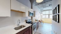 This single-floor unit blends contemporary design with industrial loft aesthetics, showcasing polished concrete floors and exposed ceiling elements throughout. The open kitchen is well-appointed with dark wood cabinetry, white quartz countertops, stainless steel appliances including a refrigerator, dishwasher, electric range, and built-in microwave, along with ample upper cabinet storage. A window at the end of the kitchen draws in natural light and frames an urban view, seamlessly connecting to a compact dining area outfitted with a wall-mounted HVAC unit.

The space includes two bedrooms: a primary room with soaring ceilings and bold dark-framed doors, and a secondary bedroom with a window offering additional natural light. The bathroom features a full tub-shower combination with clean white surroundings and chrome fixtures. A dedicated laundry room houses an in-unit washer and includes built-in shelving for added storage. Throughout, the design balances utilitarian industrial elements with modern finishes, creating a cohesive and functional urban living environment.