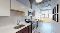 This single-floor unit blends contemporary design with industrial loft aesthetics, showcasing polished concrete floors and exposed ceiling elements throughout. The open kitchen is well-appointed with dark wood cabinetry, white quartz countertops, stainless steel appliances including a refrigerator, dishwasher, electric range, and built-in microwave, along with ample upper cabinet storage. A window at the end of the kitchen draws in natural light and frames an urban view, seamlessly connecting to a compact dining area outfitted with a wall-mounted HVAC unit.

The space includes two bedrooms: a primary room with soaring ceilings and bold dark-framed doors, and a secondary bedroom with a window offering additional natural light. The bathroom features a full tub-shower combination with clean white surroundings and chrome fixtures. A dedicated laundry room houses an in-unit washer and includes built-in shelving for added storage. Throughout, the design balances utilitarian industrial elements with modern finishes, creating a cohesive and functional urban living environment.
