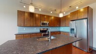 Located at 1020 N High St in Columbus, Ohio's vibrant Short North Arts District, this single-floor residence offers a thoughtfully designed layout with contemporary finishes throughout. The kitchen is a standout feature, showcasing warm medium-toned wood cabinetry that extends to the ceiling, dark granite countertops, a light blue subway tile backsplash, and a full suite of stainless steel appliances including a range, microwave, and French door refrigerator. A large central island with an undermount sink and matching granite surface provides additional prep and dining space. Track lighting and pendant fixtures illuminate the kitchen, which flows seamlessly over rich hardwood flooring into the main living area. Natural light enters through multiple large windows with clean gray framing. The bedroom offers soft wall-to-wall carpeting, a sizable window with exterior views, and a calm, neutral palette. The primary bathroom features dark espresso cabinetry, light granite countertops, an undermount sink, large mirror, and tile flooring, with access to an adjacent walk-in closet. A secondary half-bath includes a pedestal sink, hardwood flooring, and a framed mirror, offering clean, functional design. Together, these spaces create a modern and cohesive living environment.