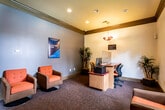 This single-room business center is designed to provide a functional and comfortable workspace for its users. The room features a warm, inviting aesthetic with neutral-toned walls accented by rich dark crown molding and baseboards, complemented by plush, multi-toned carpeted flooring throughout.

At the heart of the workspace sits a curved, red-toned granite-surfaced desk equipped with multiple computer workstations separated by clear acrylic privacy dividers, along with ergonomic task chairs and a dedicated printer station. A distinctive amber-hued pendant light fixture hangs above the workstation area, adding a warm, decorative touch to the functional setup.

A seating lounge area occupies one side of the room, furnished with retro-style tufted swivel armchairs in a bold rust-orange upholstery, ideal for casual meetings or relaxed work sessions. A large, dark-framed picture window floods this area with natural light and offers a pleasant view of a lush, tree-lined outdoor courtyard area. A wall-mounted flat-screen TV provides additional utility for presentations or media viewing.

Tall, leafy potted plants are strategically placed throughout the room, adding a refreshing touch of greenery to the professional environment. Community WiFi service is available within the space, making it a fully connected and well-rounded amenity center.