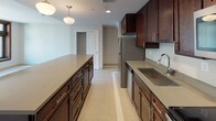 This studio unit offers a thoughtfully designed single-floor layout that maximizes its comfortable footprint. The open living area is anchored by large, dark wood-framed windows that frame urban city views and invite generous natural light, creating an airy, welcoming atmosphere throughout the space.

The kitchen is a standout feature, showcasing rich dark wood cabinetry extending to the upper walls, complemented by neutral gray countertops. A substantial kitchen island provides additional prep and storage space, while pendant lighting adds a refined touch overhead. Stainless steel appliances, including a full-size refrigerator, microwave, and range, complete the functional and stylish cooking space.

Light-toned tile flooring runs consistently throughout the unit, lending a clean, cohesive look. The bathroom is well-appointed with a dark wood vanity topped with a gray countertop and undermount sink, a wall-mounted mirror with overhead lighting, and a glass-enclosed shower. Decorative mosaic tile detailing on the bathroom floor adds a subtle, elegant design element.

Overall, the unit presents a modern, neutral aesthetic with quality finishes, practical storage, and a bright, open feel suitable for urban living.