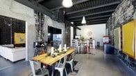 This single-floor space showcases a compelling industrial-chic aesthetic that blends raw architectural elements with carefully curated furnishings. Exposed brick and weathered stone walls form a striking backdrop, while dark-painted structural beams and visible ductwork running across the ceiling reinforce the space's loft-inspired character. The polished concrete flooring ties the design together, offering a sleek, durable surface that complements the overall industrial tone.

Lighting plays a key role in defining the ambiance. Large vintage-style pendant lights hang from the ceiling, casting a warm, inviting glow throughout the open-plan layout. The space is divided into distinct seating zones: a communal dining area anchors the foreground, featuring a reclaimed wood table surrounded by eclectic metal chairs and adorned with a botanical centerpiece. Further back, bar-height tables and stools create a more casual lounge setting near a clean, white counter area equipped for refreshments.

The open layout allows for flexible use, making it well-suited for private events, team gatherings, or social functions. The interplay of rough textures and refined details lends the space a warm yet edgy character throughout.