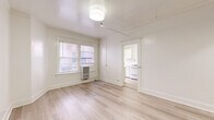 This thoughtfully laid-out one-bedroom, one-bathroom unit offers an efficient and bright living environment across a single floor. The main living area is generously proportioned relative to the overall footprint, featuring light-toned hardwood flooring, crisp white walls, and a ceiling-mounted light fixture complemented by a track rail system. A wall-mounted air conditioning unit and multiple windows allow for natural light and ventilation, with views to the surrounding neighborhood streetscape.

The galley-style kitchen is well-appointed with white cabinetry lining both sides, light stone countertops, a gas range, a refrigerator, and a single basin sink — all beneath a window that brings in additional natural light. The layout maximizes storage and counter space within a compact footprint.

The spacious bedroom includes two notable storage features: a built-in shelved closet accessible through a paneled door, and a set of sliding mirrored closet doors, adding both functionality and a sense of depth to the room. Hardwood flooring continues throughout.

The bathroom features a classic freestanding clawfoot bathtub with chrome fixtures, lending a timeless, vintage character to the space. A hallway connects the rooms, contributing to a logical and flowing floor plan. Throughout the unit, the consistent use of white tones and natural wood flooring creates a clean, cohesive, and bright aesthetic.