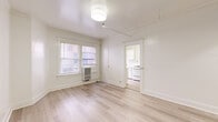 This thoughtfully laid-out one-bedroom, one-bathroom unit offers an efficient and bright living environment across a single floor. The main living area is generously proportioned relative to the overall footprint, featuring light-toned hardwood flooring, crisp white walls, and a ceiling-mounted light fixture complemented by a track rail system. A wall-mounted air conditioning unit and multiple windows allow for natural light and ventilation, with views to the surrounding neighborhood streetscape.

The galley-style kitchen is well-appointed with white cabinetry lining both sides, light stone countertops, a gas range, a refrigerator, and a single basin sink — all beneath a window that brings in additional natural light. The layout maximizes storage and counter space within a compact footprint.

The spacious bedroom includes two notable storage features: a built-in shelved closet accessible through a paneled door, and a set of sliding mirrored closet doors, adding both functionality and a sense of depth to the room. Hardwood flooring continues throughout.

The bathroom features a classic freestanding clawfoot bathtub with chrome fixtures, lending a timeless, vintage character to the space. A hallway connects the rooms, contributing to a logical and flowing floor plan. Throughout the unit, the consistent use of white tones and natural wood flooring creates a clean, cohesive, and bright aesthetic.