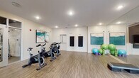 Located at 9015 Element Cir in Garner, NC, this fitness center is organized into distinct zones designed to support a wide range of workout routines. The facility is laid out across a single floor and divided into interconnected areas, each tailored to a specific type of training.

The main strength and cardio area features a dramatic dark ceiling with industrial-style pendant lighting, creating a focused atmosphere. True brand treadmills, ellipticals, and a full array of cable and resistance machines are arranged throughout, alongside a well-stocked dumbbell rack and weight benches. Colorful round artwork accents the walls, adding visual interest to the space.

Adjacent to the main floor, a bright studio area is finished with wood-style flooring and large mirrors spanning one wall. A mounted flat-screen television, step platforms, exercise balls, yoga mats, and foam rollers make it well-suited for group fitness classes or individual flexibility and balance training. Recessed lighting keeps the room evenly lit.

A second studio space features natural light from multiple windows, wood-style flooring, medicine balls, stability balls, yoga blocks, and mats — offering an open, airy environment for stretching or low-impact workouts. Throughout the facility, contemporary teal-toned artwork ties the spaces together, contributing to a clean and modern aesthetic.