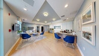 The Rumney Flats leasing office is a thoughtfully designed, single-floor commercial space that combines professional functionality with an inviting atmosphere. Upon entering through the glass-paneled front doors, visitors are welcomed into an open reception and lounge area bathed in natural light from large windows overlooking the exterior grounds. The space features light hardwood flooring throughout, soft blue-gray accent walls, and a white tray ceiling with warm flush-mount lighting that adds architectural dimension to the main area.

The reception zone is anchored by a sleek wood-finish desk accompanied by bold cobalt-blue accent chairs, establishing a consistent, contemporary color palette carried throughout. A built-in counter with bar-height seating beneath the illuminated Rumney Flats signage serves as an informal meeting spot, while a cozy lounge area with a neutral sofa, patterned area rug, and a round upholstered ottoman provides a comfortable space for prospective residents.

Two private offices branch off from the main area, each equipped with L-shaped wood desks, ergonomic seating, and coordinating blue guest chairs. Property artwork and community photography adorn the walls, reinforcing the Rumney Flats brand identity. The overall ambiance is clean, modern, and welcoming, balancing a professional leasing environment with the warmth of thoughtful interior design.