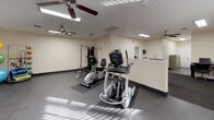 This fitness center is a comprehensively equipped exercise facility designed to support a full range of workout routines. The space is laid out across a single floor and organized to accommodate both cardio training and strength work comfortably. The flooring throughout is a durable diamond-plate rubber surface, providing excellent traction and a functional, gym-appropriate aesthetic.

The cardio section features multiple treadmills and a recumbent stationary bike, all from reputable brands including True Fitness, as well as an elliptical trainer and a stair climber, offering users a wide selection of low- and high-impact exercise options. A wall-mounted flat-screen television is positioned for easy viewing during workouts.

For strength training, the facility includes a Hoist multi-functional cable machine paired with a dedicated weight bench, allowing for a variety of resistance exercises. Stability balls in assorted sizes are neatly stored on a rack, supporting core and flexibility training.

Multiple ceiling fans are installed throughout the space to promote air circulation and comfort during exercise. Recessed fluorescent lighting provides bright, even illumination. Windows along one wall allow natural light to filter in. A large wall mirror enhances the sense of space and allows users to monitor their form. A small utility area with a sink is also accessible within the facility.
