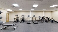 Located at 3215 Stuart Ave in Richmond, Virginia, this well-equipped fitness center offers a comprehensive single-floor exercise space designed to support a wide range of workout routines. The room features a suspended acoustic tile ceiling with recessed panel lighting that provides bright, even illumination throughout the space. The walls are finished in a neutral cream tone, contributing to a clean and functional atmosphere.

The cardio section is lined with multiple treadmills, elliptical trainers, and a stationary recumbent bike, all arranged along one wall to maximize open floor space. A dedicated rowing area houses multiple RowERG rowing machines, ideal for low-impact, full-body conditioning. For strength training, the facility includes a dual-stack functional cable trainer, an adjustable weight bench, a rack of dumbbells in varying weights, and a set of kettlebells. Wall-mounted mirrors enhance the sense of space and allow users to monitor their form during exercise.

The flooring throughout is a durable, speckled rubber material well-suited for heavy equipment and high-impact activity. A glass-paned door provides access to an adjoining area, and the layout allows for ample circulation between equipment stations. The overall design is practical and purposeful, providing residents or users with a fully functional fitness environment.