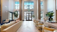 Located at 800 W Vickery Blvd in Fort Worth, TX, this single-floor amenity space serves as an impressive communal hub defined by its striking architectural design and upscale finishes. The centerpiece of the space is a grand, double-height lobby entrance flanked by floor-to-ceiling steel-framed windows that flood the interior with abundant natural light and offer open views of the surrounding courtyard and community.

The main lounge area features elegant herringbone-pattern hardwood flooring and is thoughtfully furnished with a mix of plush sofas, rounded accent chairs, and a large communal dining table with bench seating — creating distinct zones for relaxation and social gathering. A dramatic dark marble fireplace serves as a focal point, paired with custom neon wall art incorporating the "FTW 76104" zip code, giving the space a distinct local identity.

Built-in wood shelving with integrated accent lighting adds warmth and display functionality along the walls. Adjacent to the lounge is a well-appointed catering kitchen featuring light wood cabinetry, white quartz countertops, a deep undermount sink, a wall-mounted TV, and large-format porcelain tile flooring. Warm wood architectural columns and beams frame the open layout throughout, contributing to a cohesive, contemporary aesthetic that balances sophistication with comfort.