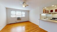 Located at 5651 N Magnolia Ave in Chicago's Edgewater neighborhood, this single-floor unit offers a clean, functional layout with consistent design elements throughout. Warm-toned hardwood flooring extends across the main living area and bedroom, lending continuity and a classic Chicago character to the space.

The open living area is bright and simply appointed, featuring white walls, a ceiling fan with light fixture, a baseboard heating system, and a wall-mounted air conditioning unit. Natural light enters through a window fitted with blinds, providing views of the surrounding neighborhood.

The kitchen is positioned adjacent to the living area and accessible via a pass-through counter, creating an open, connected feel. It is equipped with a gas range, refrigerator, under-cabinet range hood, white tile backsplash, speckled laminate countertops, and upper cabinets with wood-accented glass panels. A recessed light above the pass-through adds task lighting.

The bedroom is a comfortable, well-proportioned room with the same hardwood flooring, a ceiling fan, baseboard heat, and a window for natural light.

The bathroom features white mosaic tile flooring, a tub-shower combination with curtain, a vanity with sink, and a louvered door that opens to a built-in shelving unit, offering practical storage.